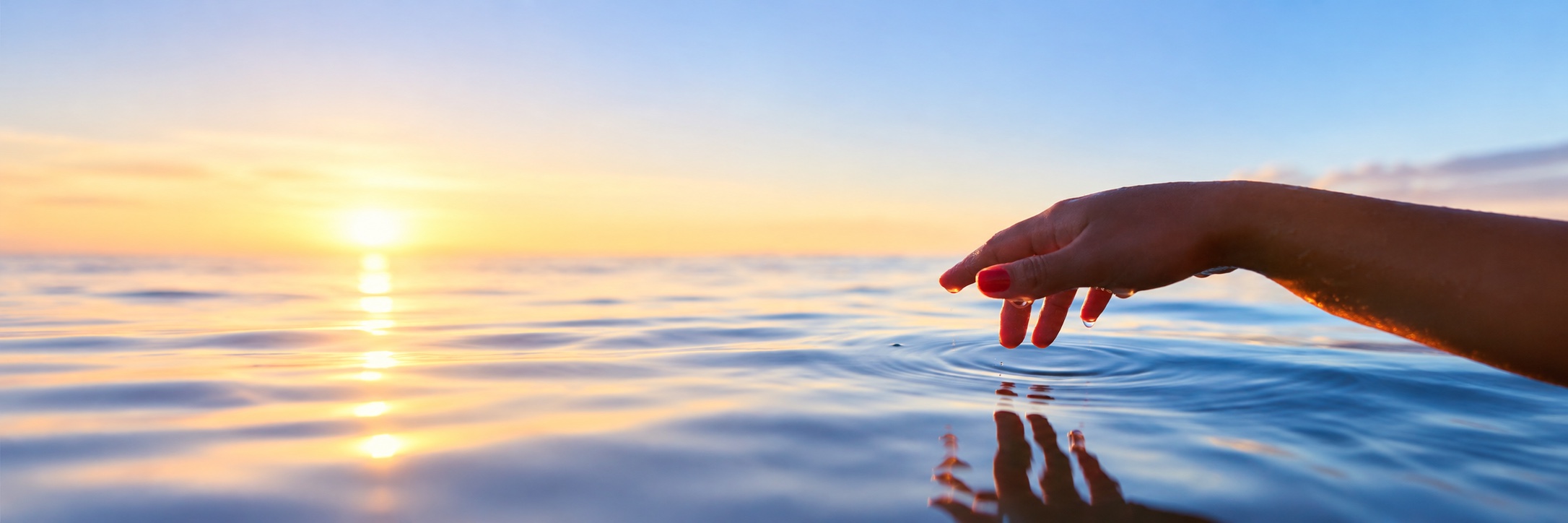 Eine Hand über ruhigem Wasser, mit Tropfen und ihrer Spiegelung im Abendlicht.